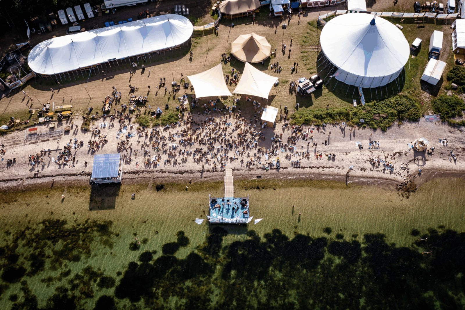 Luftfoto af Avernax Festival på Avernakø med telte, publikumsområde og scene på vandet omgivet af sol og sommerstemning.